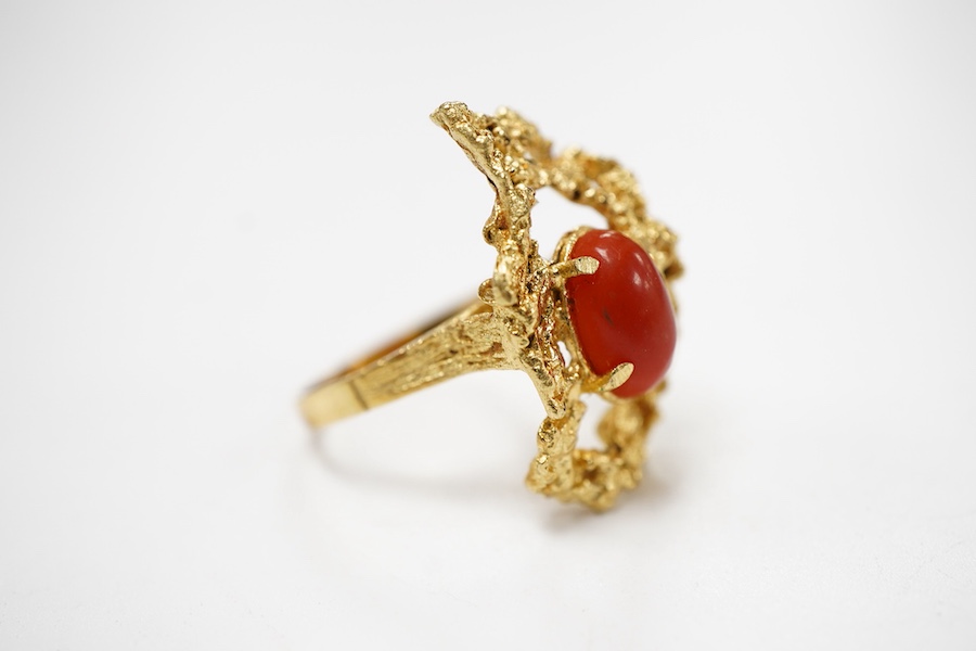 A gold and coral ring, 1970s, of textured design, set with a coral cabochon, mounted in gold, size N1/2, stamped 18K, gross weight approximately 7 grams